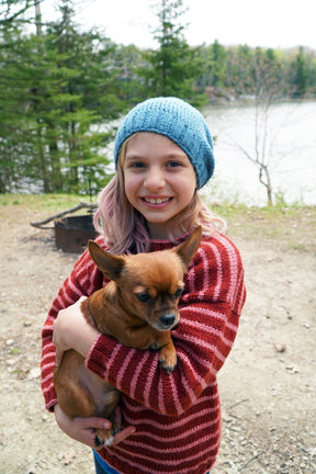 A young girl with wavy light hair, wearing a blue knit beanie and a red striped Lobster Roll Pullover by Halcyon Yarn in children's sizes, smiles while holding a small brown dog. They are outdoors near a body of water with trees in the background.