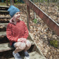 A young person with pink hair, wearing a blue knit hat and a children's-sized red striped Lobster Roll Pullover from Halcyon Yarn, sits on wooden outdoor steps during a sunny day. The background features a forest floor covered in dry leaves. They are smiling and looking into the distance.