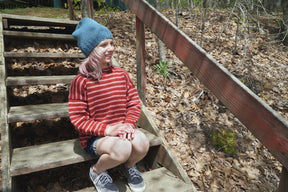 A young person with pink hair, wearing a blue knit hat and a children's-sized red striped Lobster Roll Pullover from Halcyon Yarn, sits on wooden outdoor steps during a sunny day. The background features a forest floor covered in dry leaves. They are smiling and looking into the distance.