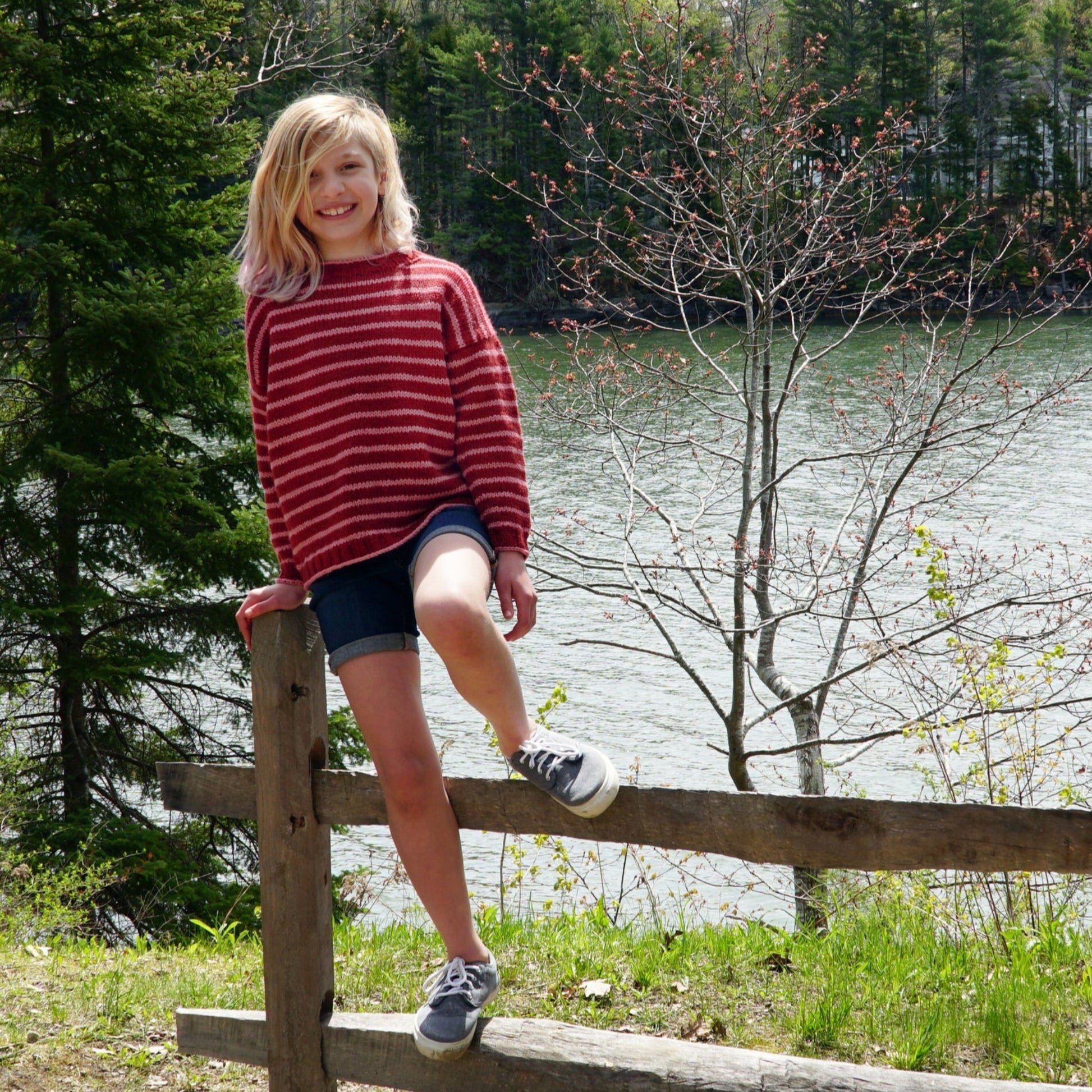 A young child with shoulder-length blonde hair, dressed in a Halcyon Yarn Lobster Roll Pullover in children's sizes and denim shorts, sits on a wooden fence. The backdrop features a tranquil lake and lush trees, suggesting a scenic, outdoor location.