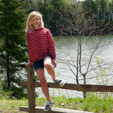 A young girl with blonde hair, wearing a Halcyon Yarn Lobster Roll Pullover in children's sizes and denim shorts, sits on a wooden fence with one leg up. Behind her is a picturesque lake surrounded by tall trees and greenery. She smiles at the camera on a sunny day.