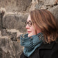 A person with shoulder-length hair, wearing glasses and a blue knitted scarf, is seen in profile. They are leaning against a textured stone wall and wearing a dark coat, perhaps on their way to share tips on making the Halcyon Yarn Saddleback Cowl using the Jacob's Ladder stitch.