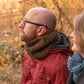 A man and woman sit closely together outdoors in an autumn setting. The man, wearing glasses and a red jacket with a knit scarf, looks up to the left. The woman, smiling and looking in the same direction, wears the Saddleback Cowl from Halcyon Yarn, which features the Jacob's Ladder stitch. Leaves and trees surround them.