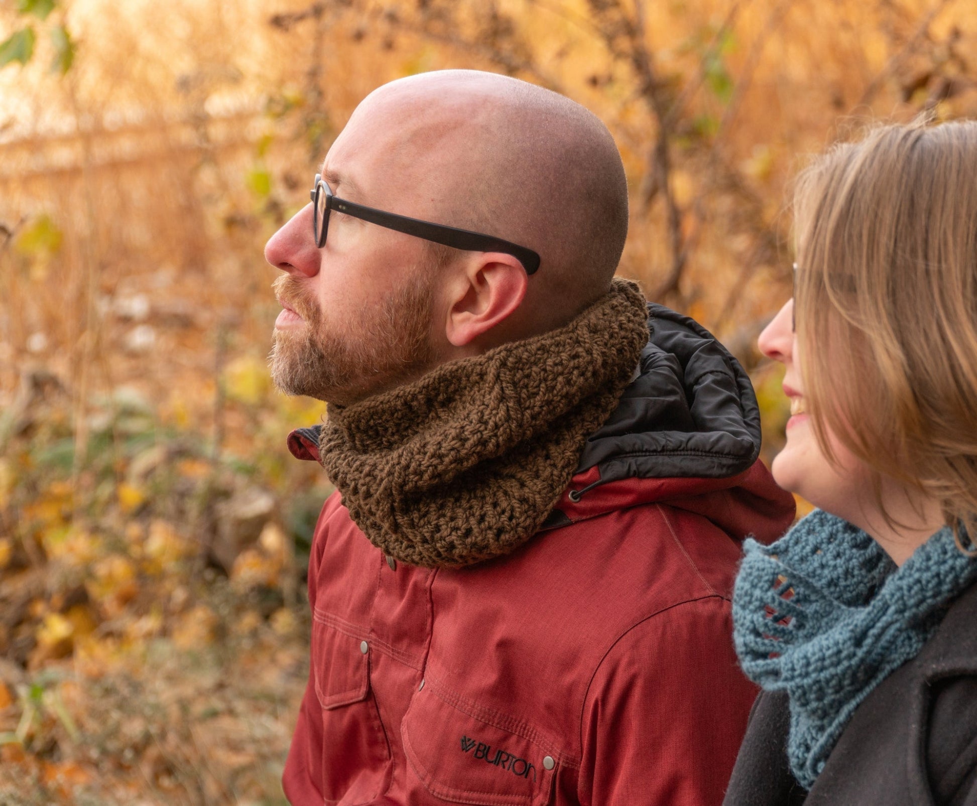 A man and woman sit closely together outdoors in an autumn setting. The man, wearing glasses and a red jacket with a knit scarf, looks up to the left. The woman, smiling and looking in the same direction, wears the Saddleback Cowl from Halcyon Yarn, which features the Jacob's Ladder stitch. Leaves and trees surround them.