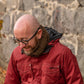 A bald man with glasses wearing a red jacket and a knitted scarf stands smiling and looking down. He is outdoors in front of a stone wall with fallen leaves scattered on the ground, perhaps admiring his recent fun and easy project featuring the Saddleback Cowl by Halcyon Yarn.