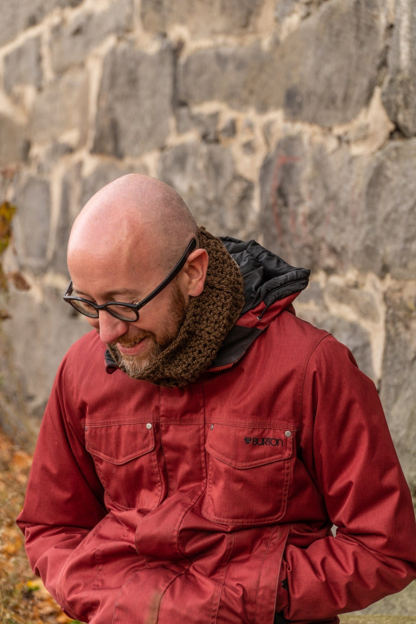 A bald man with glasses wearing a red jacket and a knitted scarf stands smiling and looking down. He is outdoors in front of a stone wall with fallen leaves scattered on the ground, perhaps admiring his recent fun and easy project featuring the Saddleback Cowl by Halcyon Yarn.