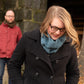 A person wearing glasses and a Halcyon Yarn Saddleback Cowl smiles and looks down while another in a red jacket with a brown scarf stands in the background. They are outdoors, near a large stone structure and a tree, surrounded by some dry foliage. The Saddleback Cowl adds warmth to the scene.