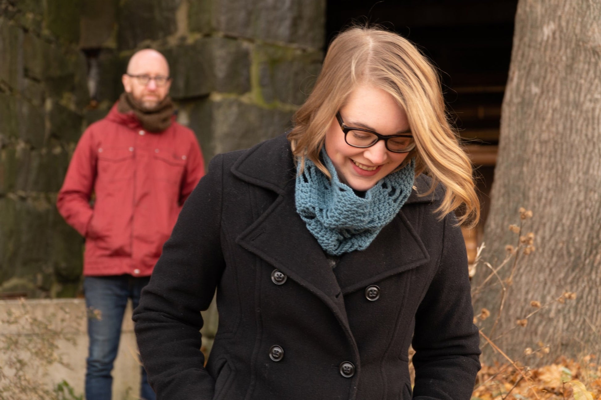 A person wearing glasses and a Halcyon Yarn Saddleback Cowl smiles and looks down while another in a red jacket with a brown scarf stands in the background. They are outdoors, near a large stone structure and a tree, surrounded by some dry foliage. The Saddleback Cowl adds warmth to the scene.