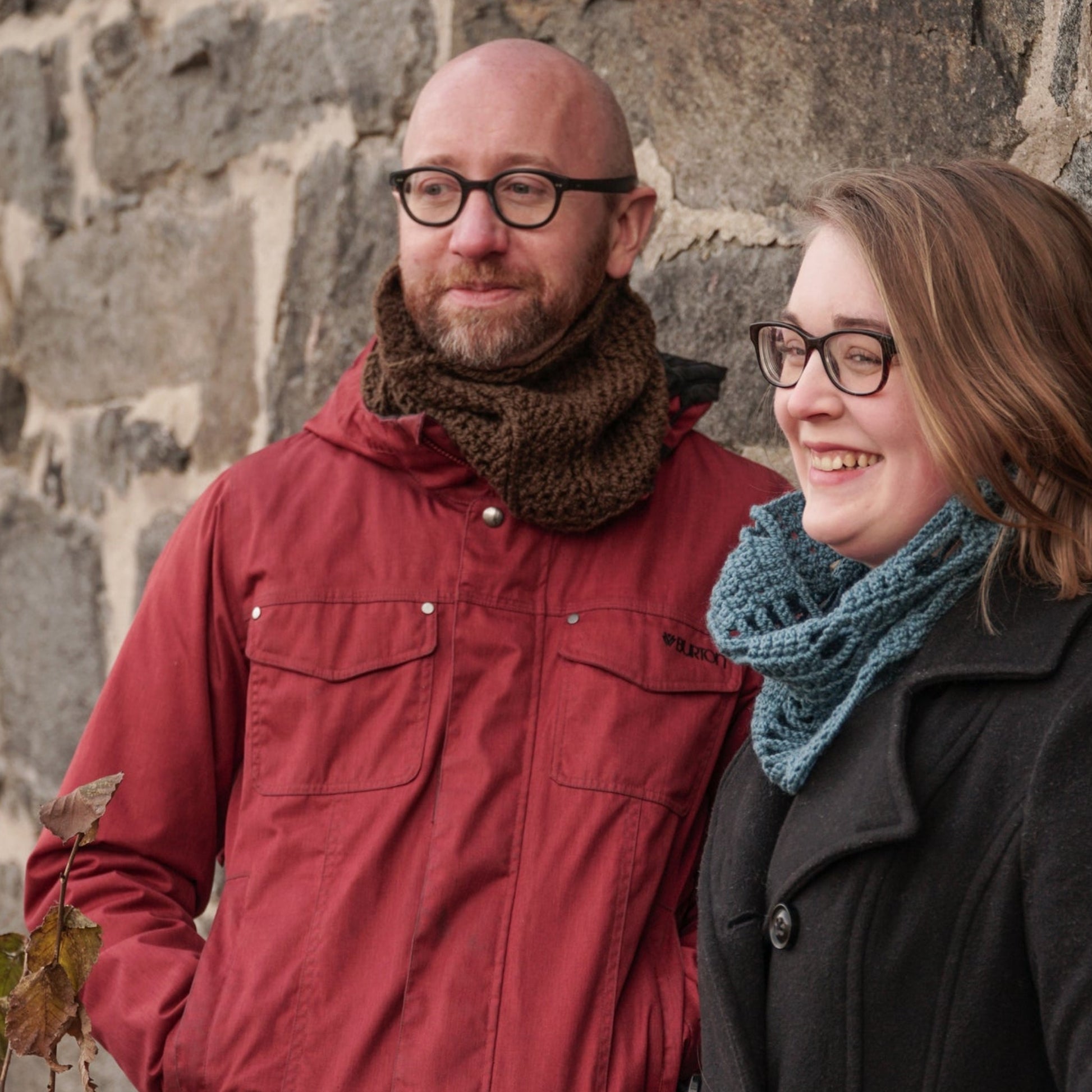 A man and a woman stand close together in front of a stone wall, each warmly wrapped in Halcyon Yarn's Saddleback Cowls. Both wear glasses and scarves, with the man sporting a red jacket paired with a brown scarf, and the woman wearing a dark coat complemented by a blue scarf. They smile warmly while looking slightly to the left.