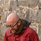 A person with a bald head, black-framed glasses, and a brown scarf wears a red jacket while standing outdoors in front of a stone wall, looking down and smiling. The brown scarf is intricately designed with Jacob's Ladder stitch patterns and is known as the Saddleback Cowl from Halcyon Yarn.