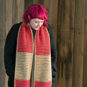 A person with bright pink hair is looking down while wearing the Lovelight Crochet Scarf by Halcyon Yarn, which is adorned with red and beige blocks and known for being large and easy to crochet. They are dressed in a dark sweater and standing against a wooden wall background. This cozy accessory, made from Aran weight yarn, adds a colorful touch to their outfit.