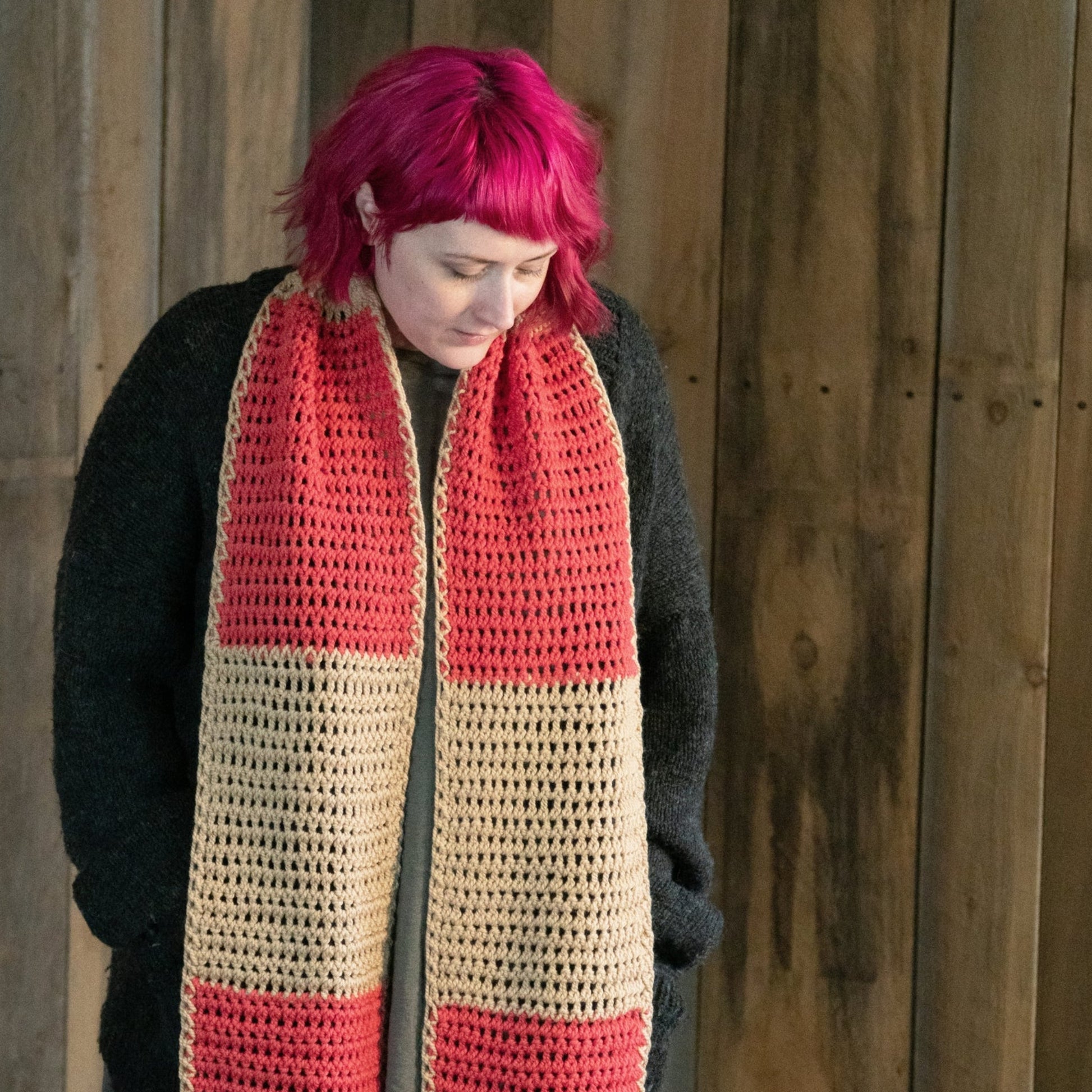 A person with vibrant pink hair, wearing a black sweater, looks down while standing against a wooden wall. They are donning the Lovelight Crochet Scarf from Halcyon Yarn, featuring wide red and beige stripes and crafted from Aran weight yarn.