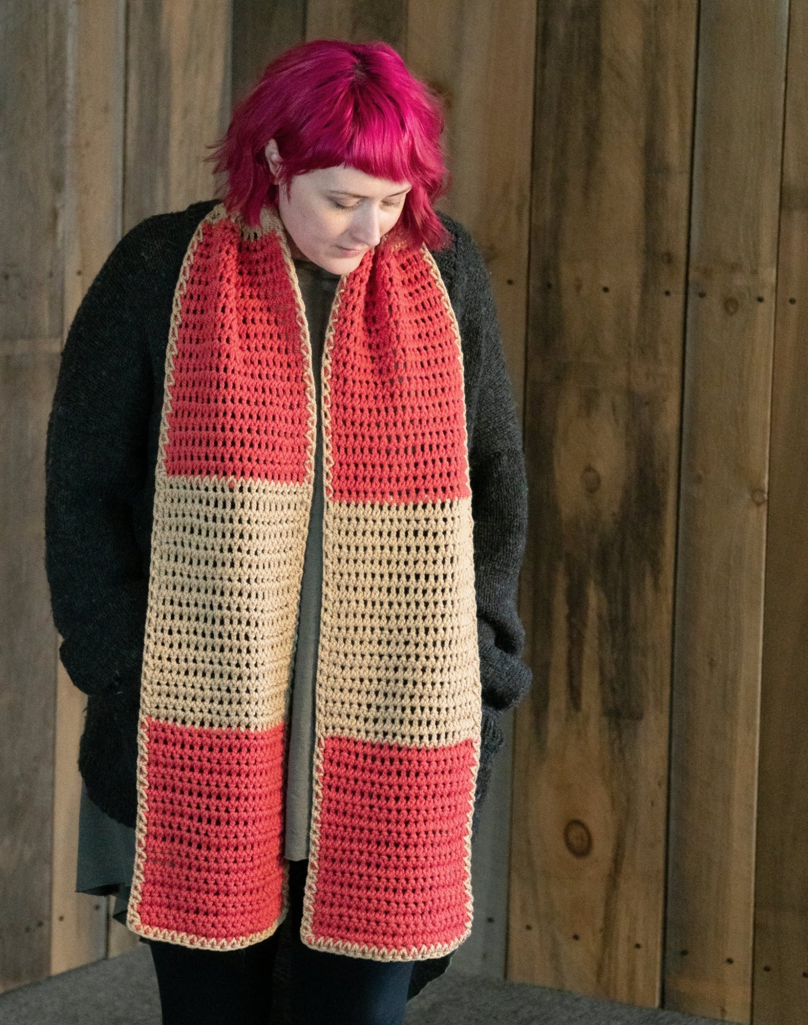 A person with pink hair wearing a dark coat and a Lovelight Crochet Scarf from Halcyon Yarn, made of Aran weight yarn featuring alternating red and beige squares, stands against a wooden paneled background, looking down.