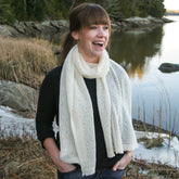 A woman with bangs, wearing a black top and the luxurious Halcyon Yarn Heirloom Lace Cashmere Wrap, stands outdoors near a calm body of water and rocky terrain. She is smiling and has her hands in her pockets. Leafs and tall grass are visible in the background.