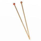 Two handpainted Peace Fleece Single-Pointed Wooden Knitting Needles, each measuring 10 inches and crafted by Harrisville Designs. The left needle is adorned with a small round, orange ornament featuring a smiling face at the end, while the right needle showcases an intricate yellow and red floral pattern. They are arranged diagonally against a white background.