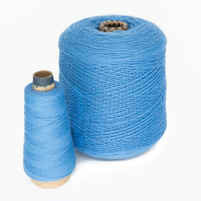 Two spools of JaggerSpun yarns from Jagger Brothers, Inc. are placed against a white background. The larger spool of JaggerSpun Maine Line 2/8 Yarn is cylindrical, while the smaller spool is cone-shaped. Both spools have threads neatly wound around them, showcasing the texture and vibrant blue color of the worsted spun wool.