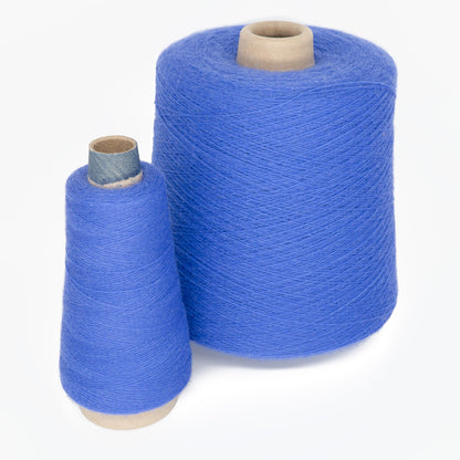 Two cones of Jagger Brothers, Inc. yarn are positioned against a plain background. The larger cone is cylindrical and wrapped with blue thread from the JaggerSpun Maine Line 2/20 Yarn | Large Cone, while the smaller cone, also cylindrical but differently shaped, rests upright on its base. Both cones are made of worsted spun yarn.
