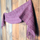 The Rivers and Roads Woven Scarf by Halcyon Yarn, featuring a plum hue and checkered pattern, is draped on a rustic wooden wall. Crafted using a weaving loom, the fringed-edged scarf hangs loosely, showcasing its detailed texture and artisanal craftsmanship against the warm-toned wood backdrop.