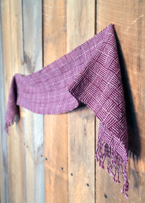 The Rivers and Roads Woven Scarf by Halcyon Yarn, featuring a plum hue and checkered pattern, is draped on a rustic wooden wall. Crafted using a weaving loom, the fringed-edged scarf hangs loosely, showcasing its detailed texture and artisanal craftsmanship against the warm-toned wood backdrop.