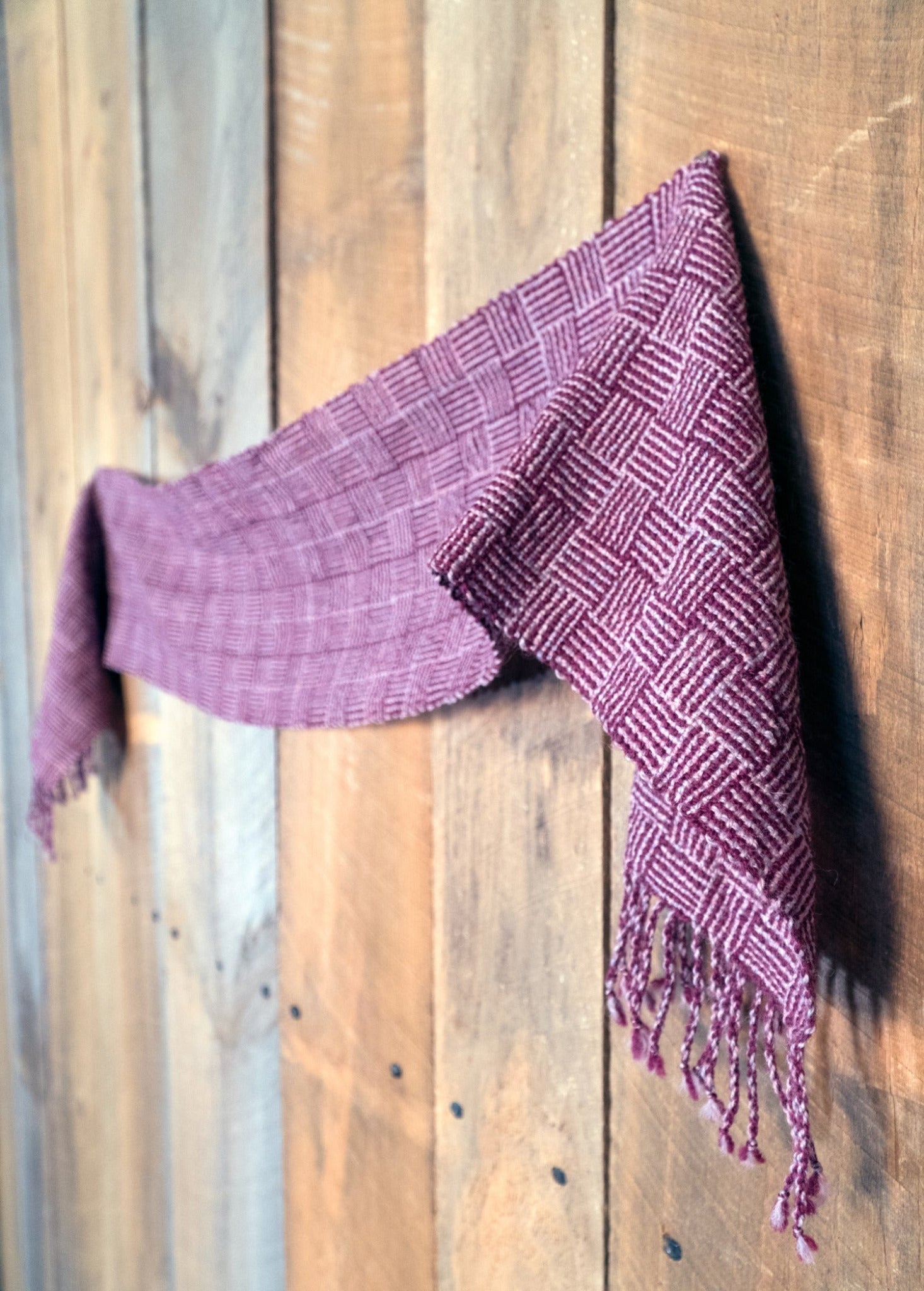 A rustic wooden wall serves as the perfect backdrop for the Rivers and Roads Woven Scarf by Halcyon Yarn, featuring a red checkered wool design with fringe. The intricate Log cabin pattern, meticulously crafted using traditional weaving techniques, adds an exquisite touch to this cozy accessory.