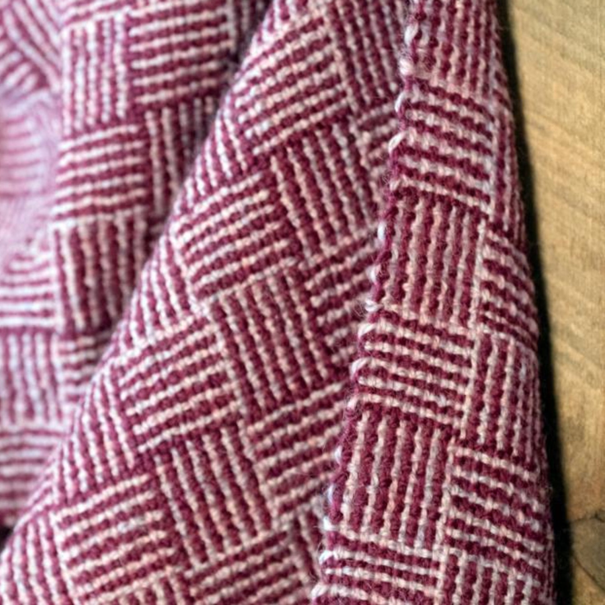 Close-up of the Rivers and Roads Woven Scarf by Halcyon Yarn, featuring a pink and white checkered fabric reminiscent of a log cabin pattern, draped over a wooden surface. The textile showcases a repeating square motif with textured, raised stitching.