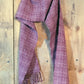 A maroon Rivers and Roads Woven Scarf by Halcyon Yarn, featuring a log cabin pattern and fringed ends, hangs against a rustic wooden wall. The scarf appears to be made of a warm, woven wool fabric.