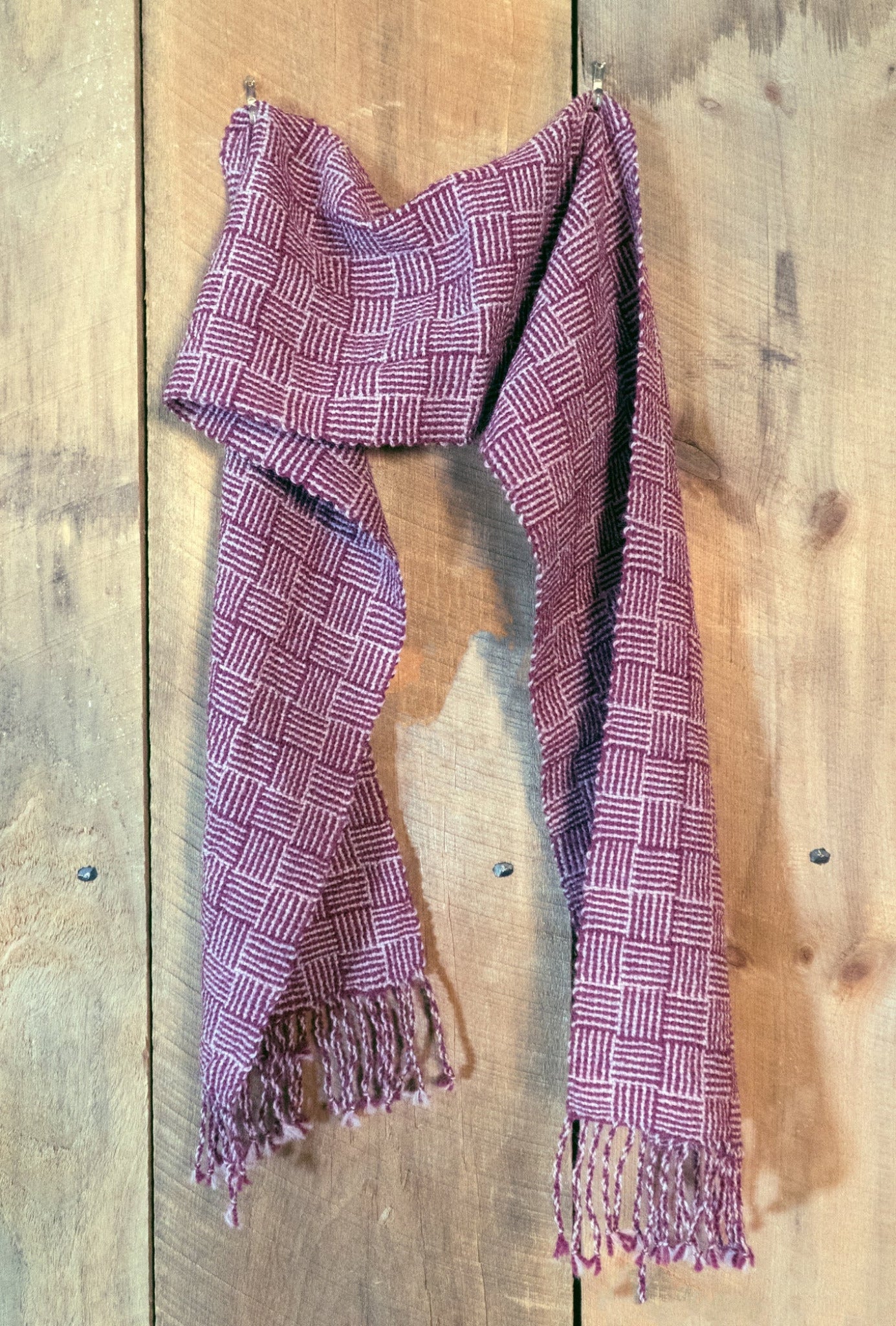 A maroon Rivers and Roads Woven Scarf by Halcyon Yarn, featuring a log cabin pattern and fringed ends, hangs against a rustic wooden wall. The scarf appears to be made of a warm, woven wool fabric.