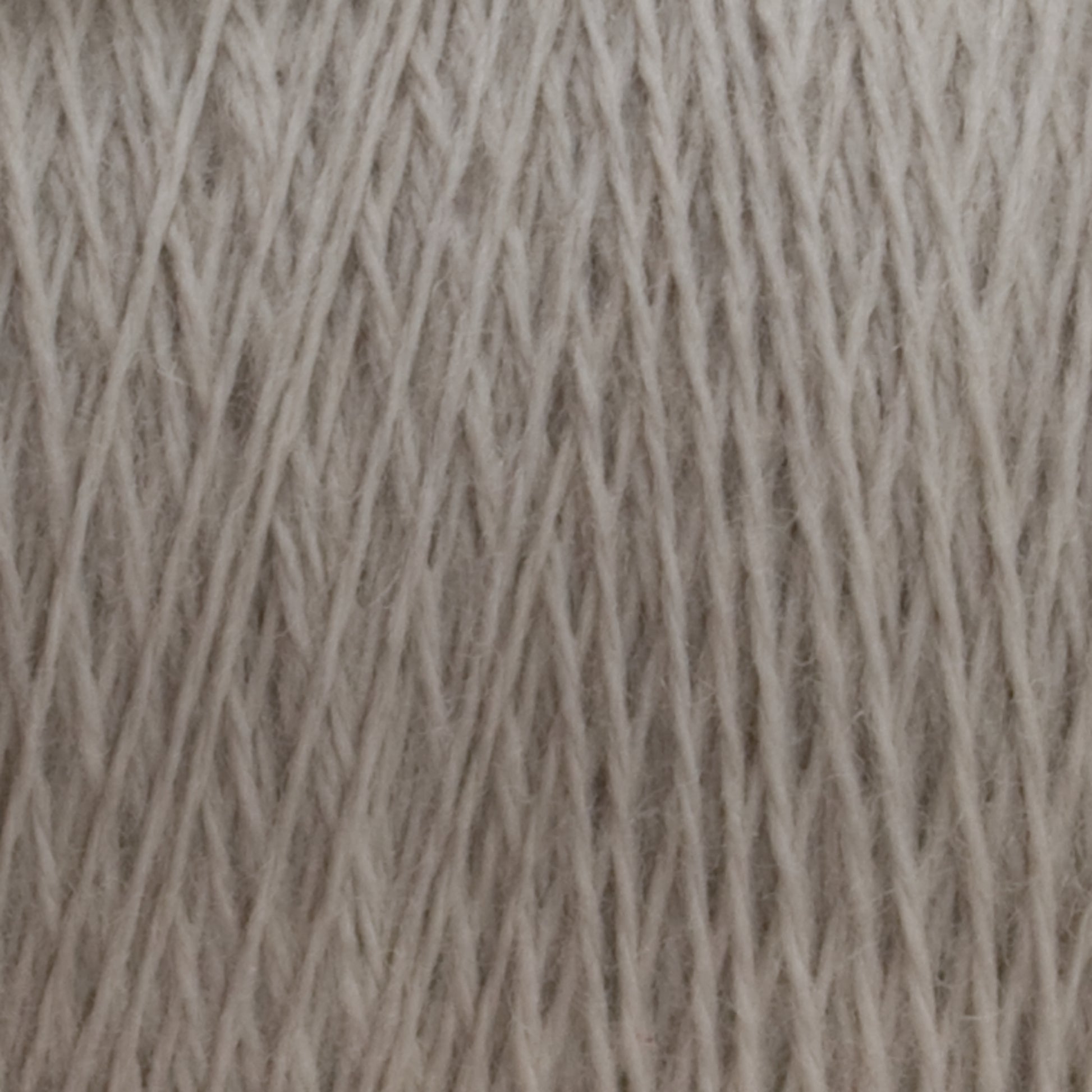 Close-up view of a large cone of beige JaggerSpun Superfine Merino 2/18 Yarn by Jagger Brothers, Inc., showcasing the detailed texture and interwoven fibers. The worsted weight yarn strands are neatly aligned, creating a pattern of diagonal lines across the image. Ideal for hand knitters seeking quality materials.