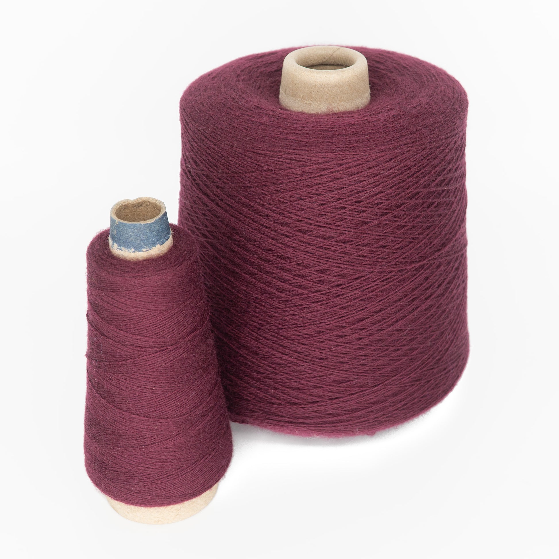 Two spools of dark red thread are pictured. The spool on the left is a smaller, tapered mini-cone, while the spool on the right is larger and cylindrical. Crafted from high-quality JaggerSpun Superfine Merino 2/18 yarn by Jagger Brothers, Inc., these spools are perfect for hand knitters. Both spools stand upright on a white background.