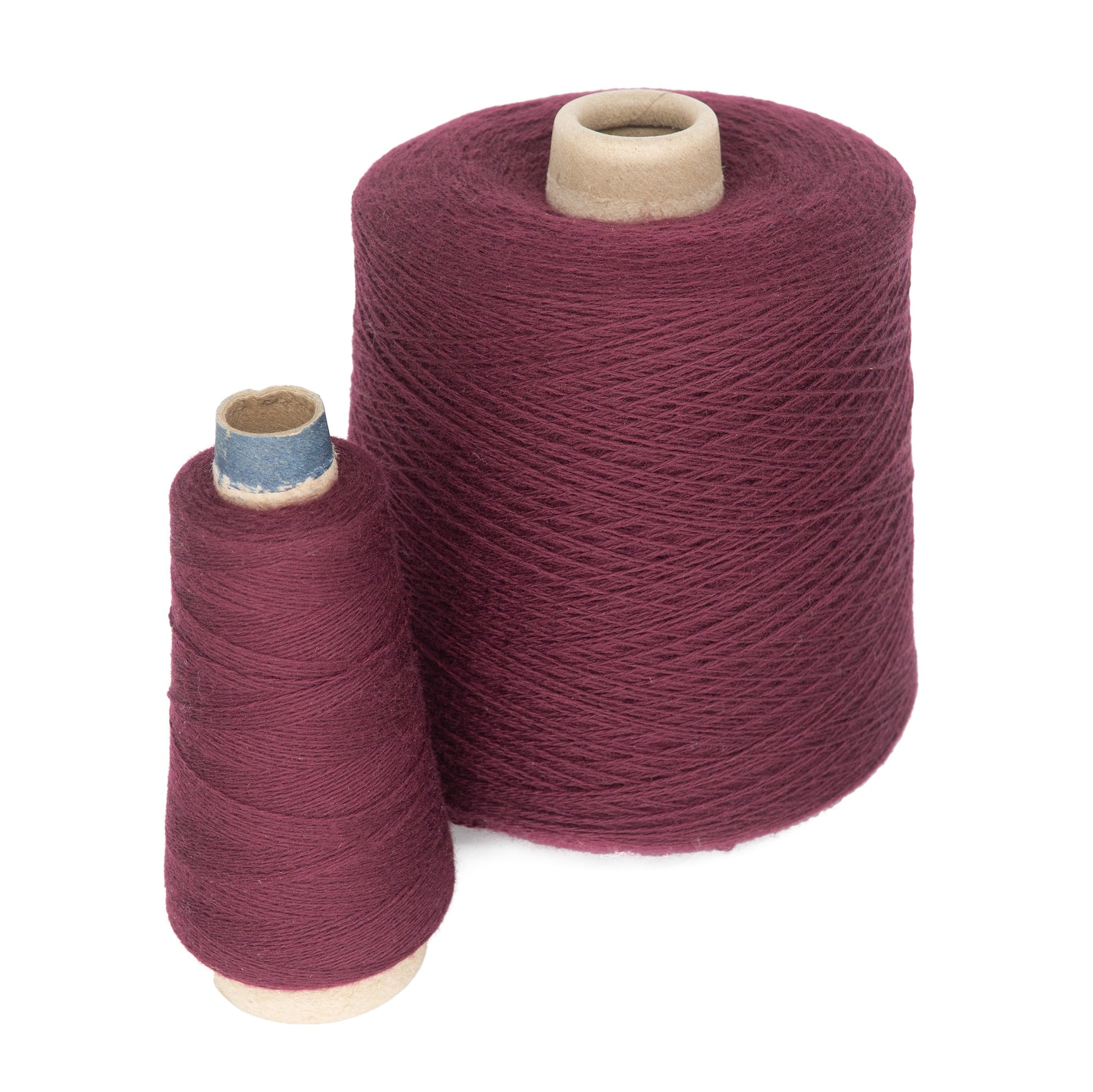 Two cones of burgundy yarn rest side by side. The larger cone, the JaggerSpun Superfine Merino 2/18 Yarn from Jagger Brothers, Inc., is positioned behind the smaller one, both made from cylindrical cardboard with tightly wound yarn, ideal for hand knitters or weavers seeking quality material for their sewing or knitting masterpieces.