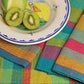 A colorful scene featuring a plate with a sliced kiwi and a partially sliced green apple. The plate has a whimsical bird illustration. Complementing the setup, vibrant, multicolored Organic Cottolin Tea Towels by Halcyon Yarn lie underneath.