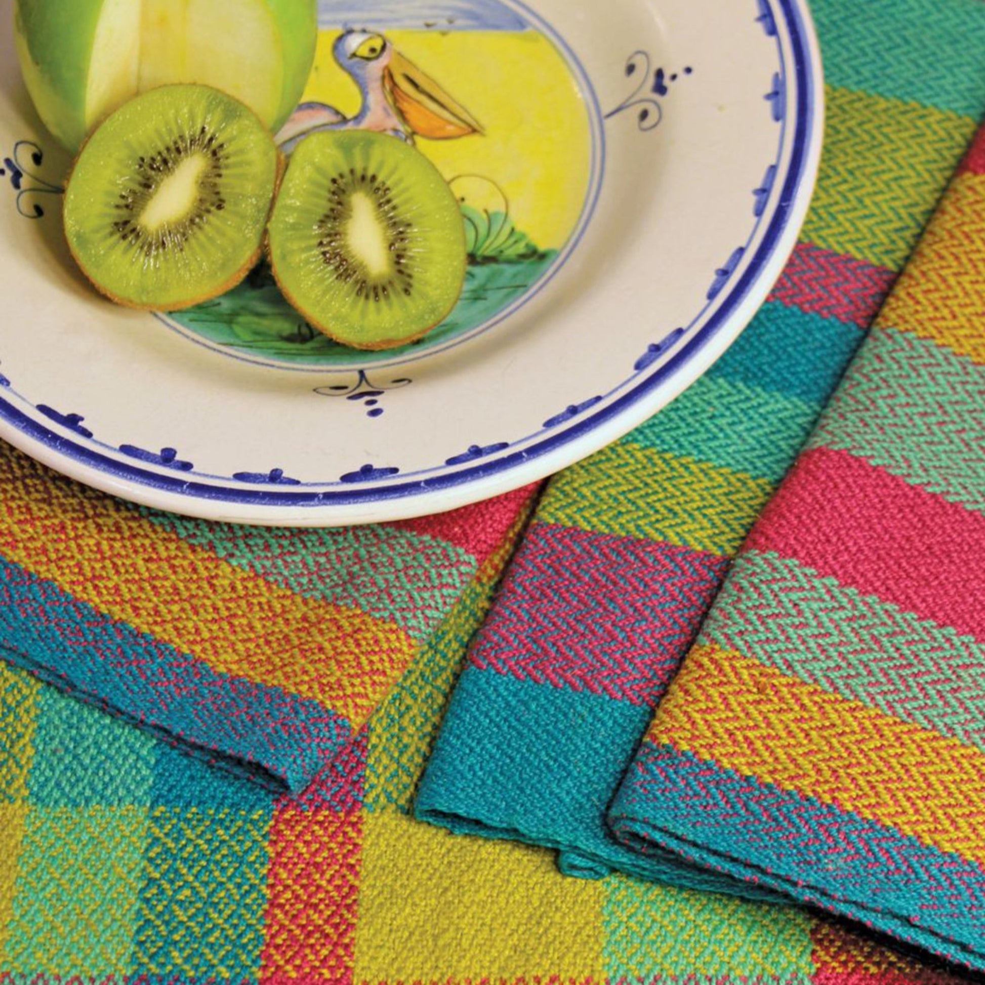 A colorful scene featuring a plate with a sliced kiwi and a partially sliced green apple. The plate has a whimsical bird illustration. Complementing the setup, vibrant, multicolored Organic Cottolin Tea Towels by Halcyon Yarn lie underneath.