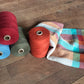 Four large spools of Halcyon Yarn organic cottolin yarn in red, blue, green, and orange are placed on a wooden floor beside a folded, colorful woven fabric in hues of orange, blue, green, and beige. The yarn is neatly wound and the fabric appears to be one of Halcyon Yarn's Organic Cottolin Tea Towels created on a 4 shaft loom.