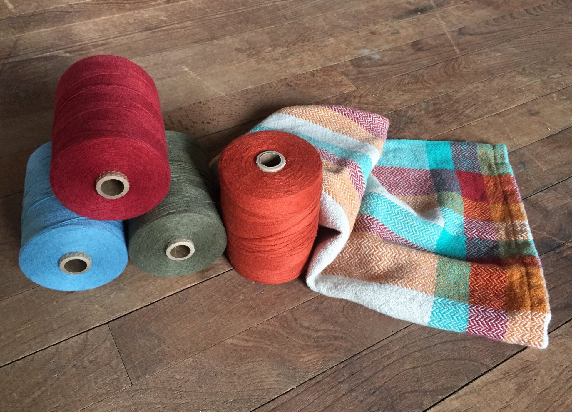 Spools of yarn in red, green, blue, and orange are arranged next to a colorful, patterned Organic Cottolin Tea Towel from Halcyon Yarn on a wooden surface. The fabric features a mix of orange, red, green, blue, and beige hues crafted from organic cottolin.