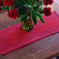 A wooden table with a Ruby Overshot Runner from Halcyon Yarn, adorned by a glass vase containing a bouquet of red roses surrounded by lush green leaves. The table is decorated with four placemats woven from organic cottolin and features engraved markings, partially visible in the cozy and inviting setting.