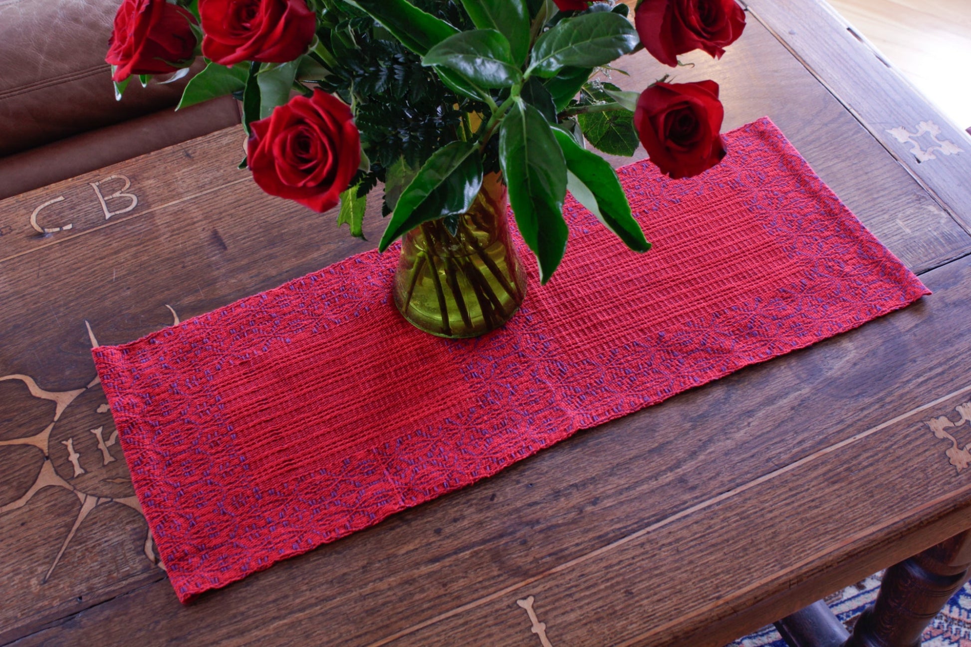 A wooden table with a Ruby Overshot Runner from Halcyon Yarn, adorned by a glass vase containing a bouquet of red roses surrounded by lush green leaves. The table is decorated with four placemats woven from organic cottolin and features engraved markings, partially visible in the cozy and inviting setting.