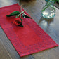 A wooden table with the Ruby Overshot Runner from Halcyon Yarn, featuring an intricate pattern in red. Accompanying the runner are four placemats made of organic cottolin. A single red rose with leaves is placed on the runner, and a glass vase with green leafy stems is positioned nearby on the table.