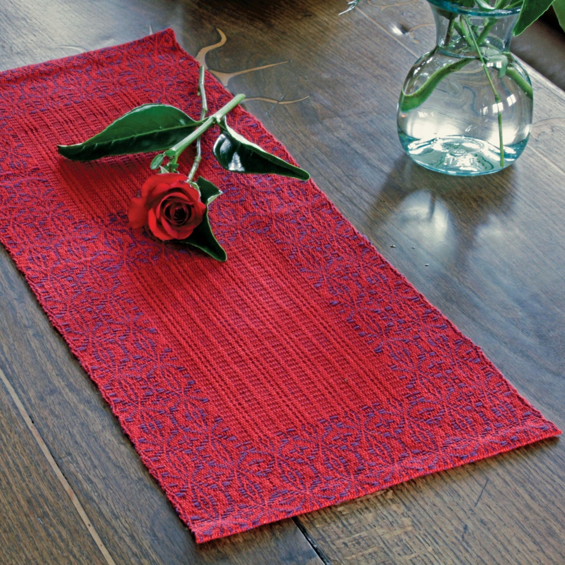 A wooden table with the Ruby Overshot Runner from Halcyon Yarn, featuring an intricate pattern in red. Accompanying the runner are four placemats made of organic cottolin. A single red rose with leaves is placed on the runner, and a glass vase with green leafy stems is positioned nearby on the table.