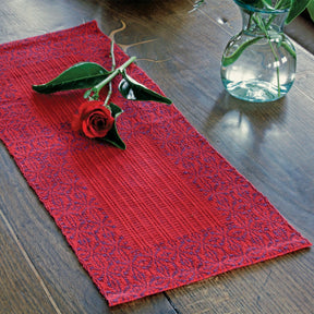 A single red rose rests on a beautifully crafted Ruby Overshot Runner, available for download from Halcyon Yarn, placed on a wooden table. Near the edge of the table, a clear glass vase with green leaves adds elegance to the Ruby Anniversary-colored setting.