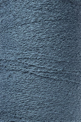 A close-up image of a spool of Maurice Brassard Cotton Bouclé Yarn. The yarn, tightly wound on a tube, exhibits visible fibers and slight irregularities that enhance its textured appearance. The muted, cool blue color of the yarn suggests a soft and cozy material ideal for creating Maurice Brassard weavings.