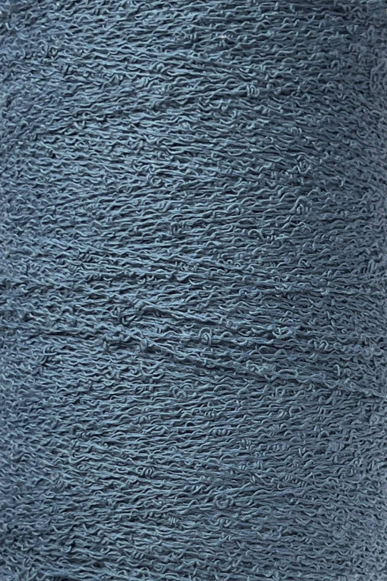 A close-up image of a spool of Maurice Brassard Cotton Bouclé Yarn. The yarn, tightly wound on a tube, exhibits visible fibers and slight irregularities that enhance its textured appearance. The muted, cool blue color of the yarn suggests a soft and cozy material ideal for creating Maurice Brassard weavings.