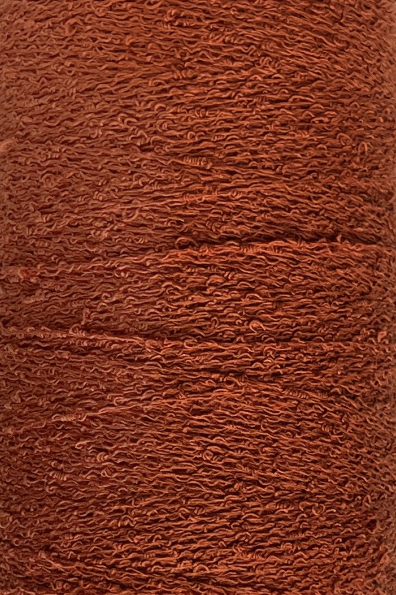 Close-up image of a spool of reddish-brown yarn with a textured, slightly wavy appearance. The densely wound Maurice Brassard Cotton Bouclé Yarn | Tube showcases intricate and uniform layers throughout.