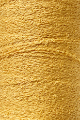 Close-up of a textured spool of Maurice Brassard's Cotton Bouclé Yarn in a golden-yellow hue, showcasing its rough and wavy fibers tightly wrapped around itself. The weaving yarn's slightly uneven surface lends it a rich, tactile quality.