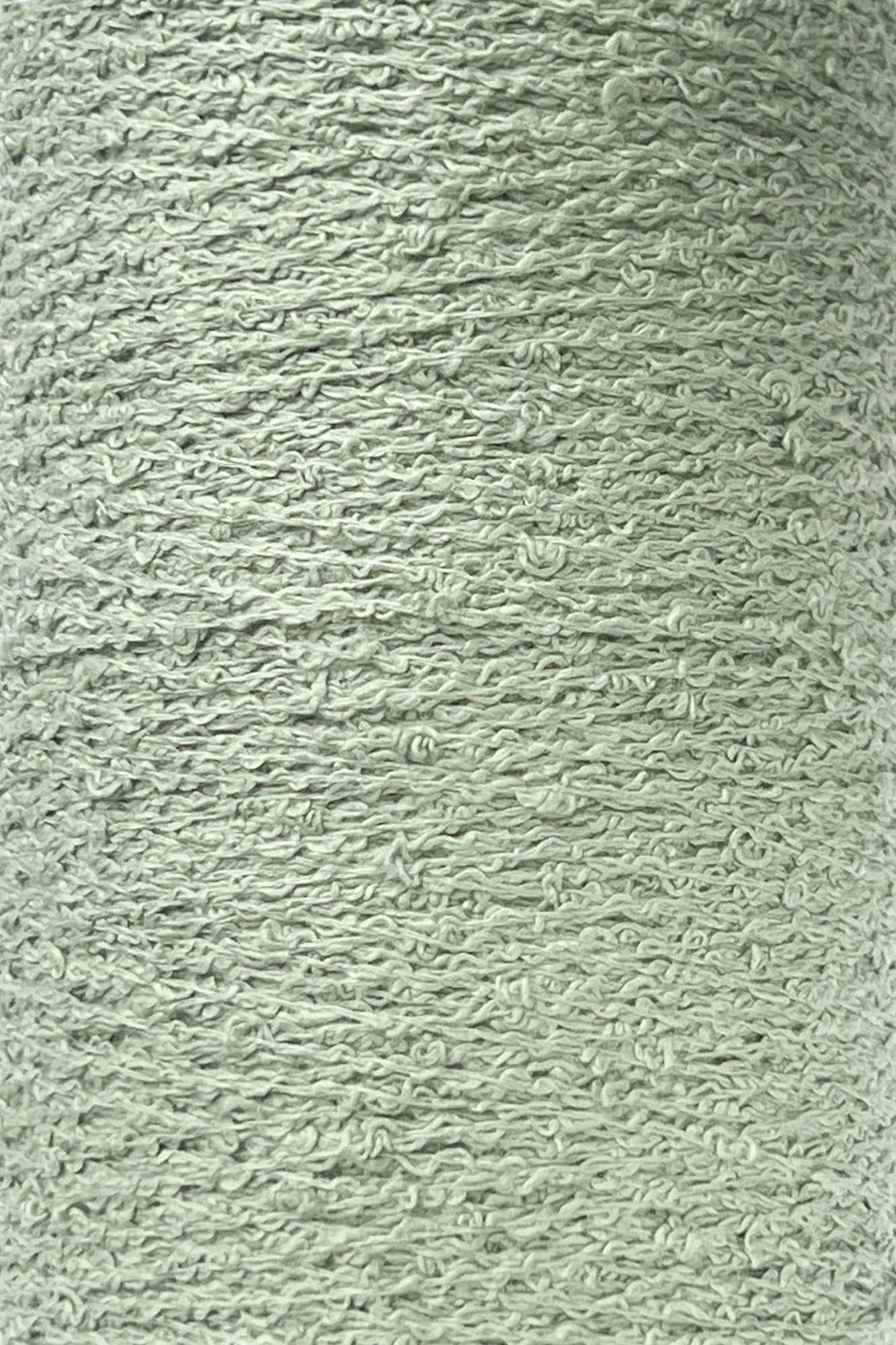 A close-up image of a spool of Maurice Brassard's Cotton Bouclé Yarn | Tube in light grey-green. The yarn displays a slightly rough and textured appearance, with visible individual fibers and small loops throughout. The color is consistently spread across the entire surface, making it ideal for various cotton weaving projects.