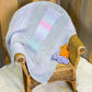 A wooden rocking chair is draped with the Dream Baby, Baby Blanket from Halcyon Yarn, offering super snuggly comfort in soft pastel colors like grey, lavender, and pink. On the chair's seat sit two small stuffed animals, one orange and one purple. The backdrop includes wooden panels and a light-colored floor.