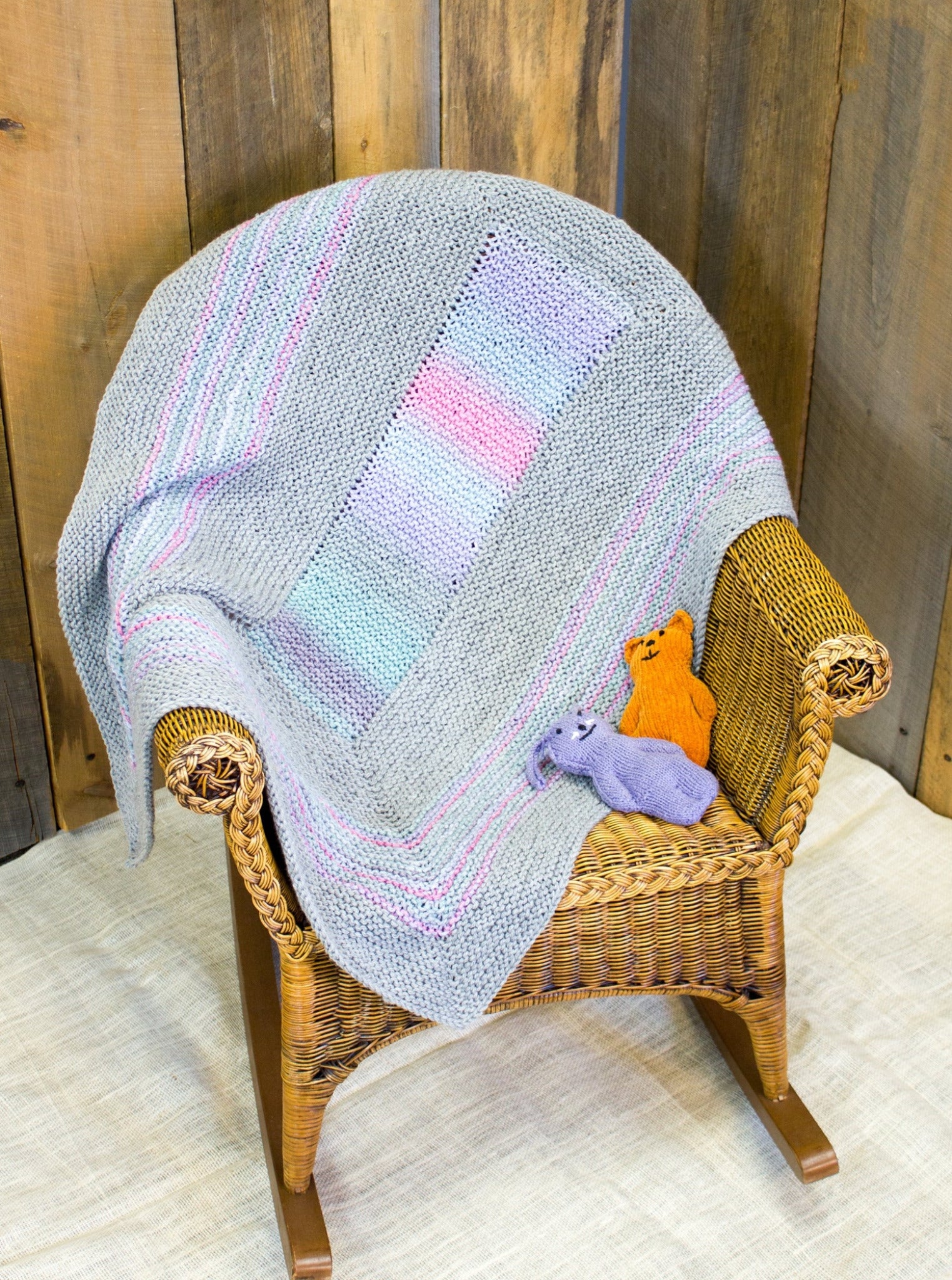 A wooden rocking chair is draped with the Dream Baby, Baby Blanket from Halcyon Yarn, offering super snuggly comfort in soft pastel colors like grey, lavender, and pink. On the chair's seat sit two small stuffed animals, one orange and one purple. The backdrop includes wooden panels and a light-colored floor.