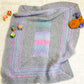 Introducing the Dream Baby, Baby Blanket by Halcyon Yarn! This ultra-cozy blanket features a delightful gray knit with a charming pastel rainbow pattern at the center and elegant striped borders. Crafted from chunky superwash yarn, it drapes beautifully over light fabric. Complete your nursery look with colorful letter and animal blocks alongside two plush toys in vibrant purple and orange hues.