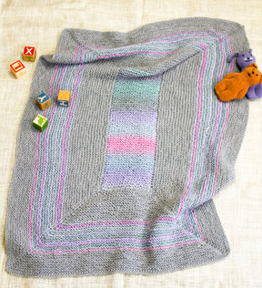 Introducing the Dream Baby, Baby Blanket by Halcyon Yarn! This ultra-cozy blanket features a delightful gray knit with a charming pastel rainbow pattern at the center and elegant striped borders. Crafted from chunky superwash yarn, it drapes beautifully over light fabric. Complete your nursery look with colorful letter and animal blocks alongside two plush toys in vibrant purple and orange hues.