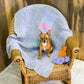 A small brown dog sits on a wicker chair draped with the super snuggly Dream Baby, Baby Blanket by Halcyon Yarn, which features pastel stripes. Two knitted stuffed animals—one resembling a yellow bear and the other a purple cat—are placed on the chair beside the dog. A wooden wall forms the background.