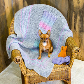 A small brown dog sits on a wicker chair draped with the super snuggly Dream Baby, Baby Blanket by Halcyon Yarn, which features pastel stripes. Two knitted stuffed animals—one resembling a yellow bear and the other a purple cat—are placed on the chair beside the dog. A wooden wall forms the background.